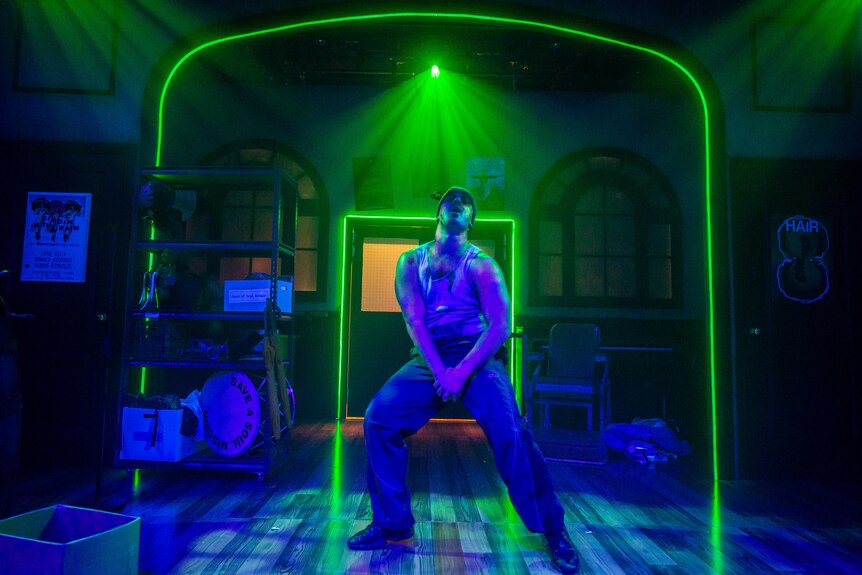 A Latino Australian man in his 30s dressed in streetwear with zombie make-up dances on a stage, backlit with green neon lights.