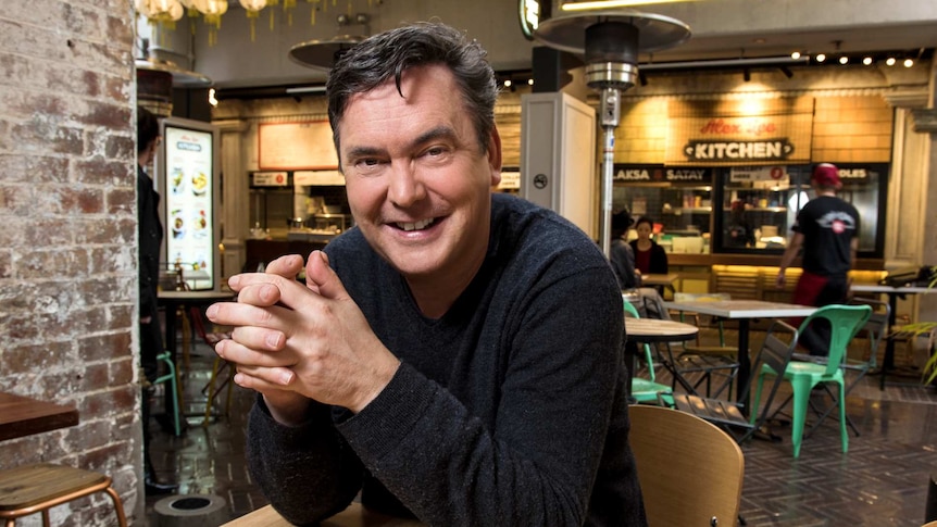 A man with short dark hair, wearing a dark jumper, sitting in a cafe.