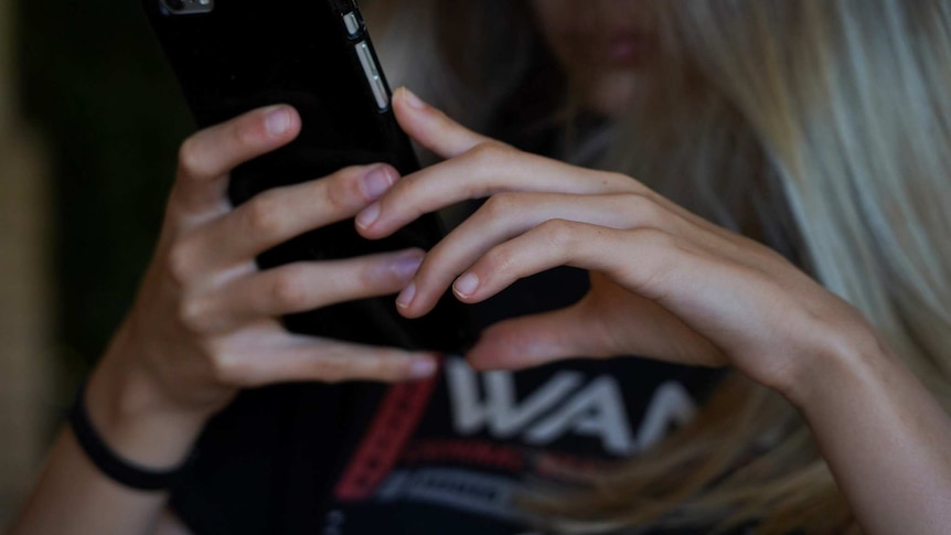 A teenage girl with blonde hair holds a mobile phone with both hands, with the phone in focus and her face obscured.