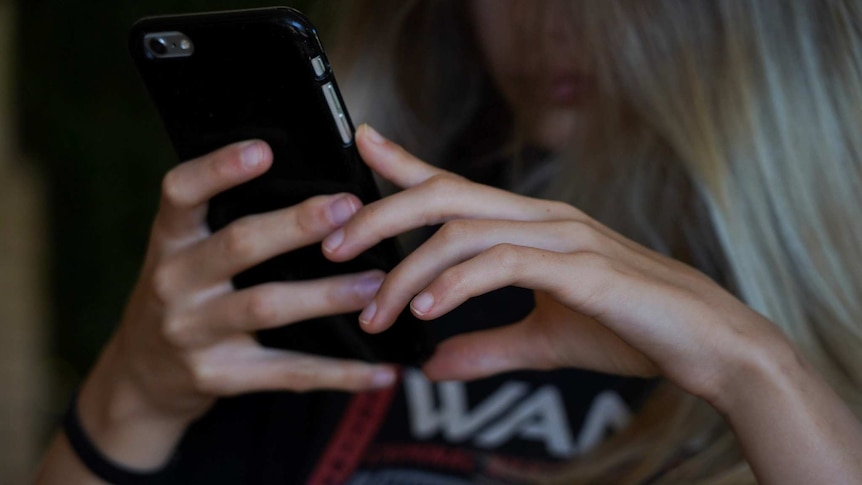 A teenage girl with blonde hair holds a mobile phone with both hands, with the phone in focus and her face obscured.