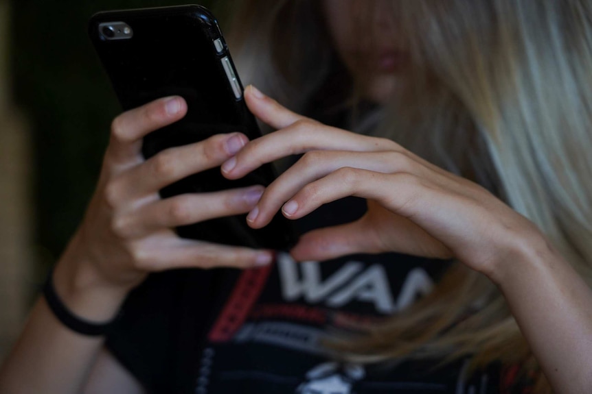 A teenage girl with blonde hair holds a mobile phone with both hands, with the phone in focus and her face obscured.