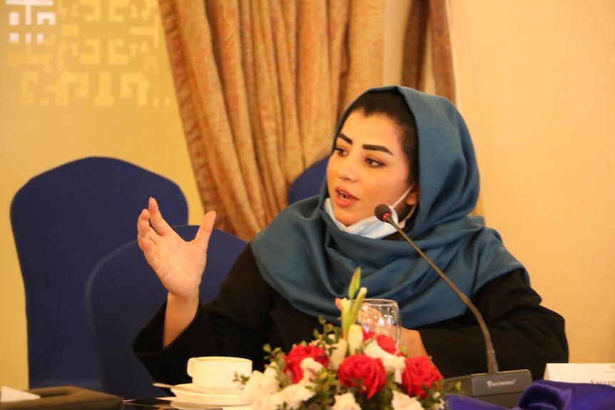 A women wearing a blue headscarf speaks into a microphone from behind a table.