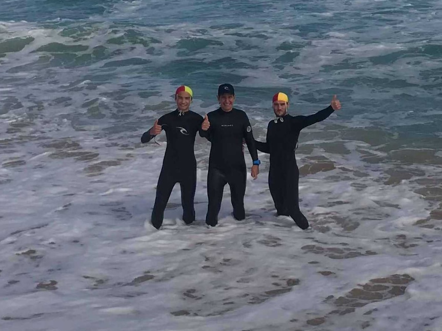 Three people in wet suits wave from within the surf where they are standing at knee-depth.