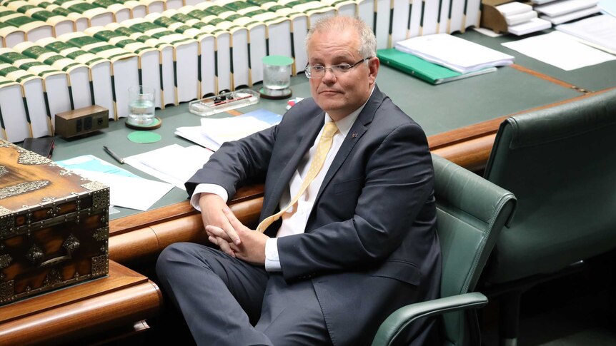 Morrison sits in his chair looking contemplative. Legs crossed, hands together in lap.