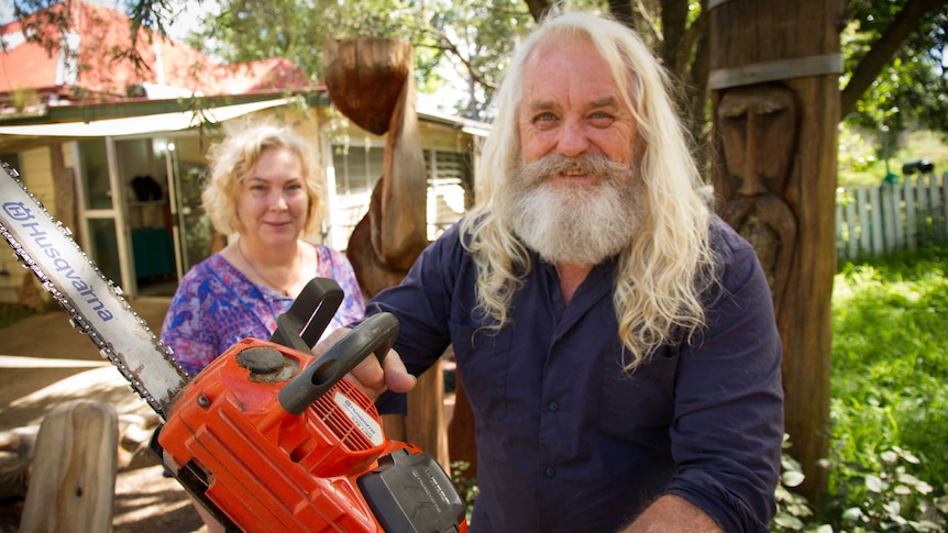 A long haired grey-bearded smiling man holds a chainsaw near a totem, while his wife, also an artist, looks on.