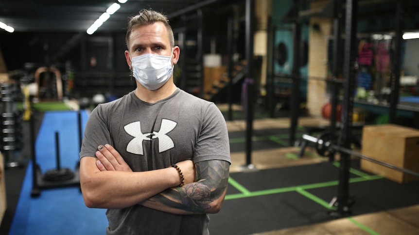 A man with tattoos and a face mask stands in an empty gym