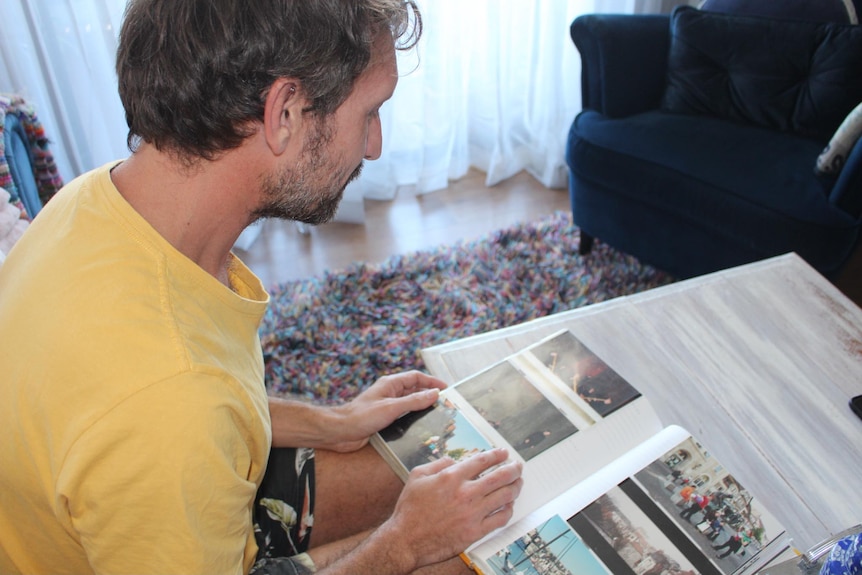 Man in yellow shirt sitting on couch flicks through photo album of childhood photos.