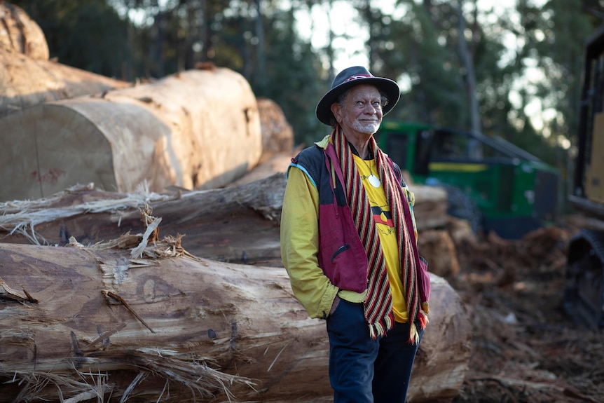 A man wearing Aboriginal colours and wide brim hat stands and smiles at camera with pile of logs behind him