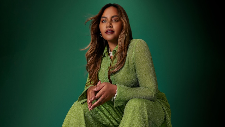 Jessica Mauboy wearing a light green jumpsuit sitting down against a dark green background, long hair, hands together on knees