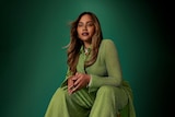 Jessica Mauboy wearing a light green jumpsuit sitting down against a dark green background, long hair, hands together on knees