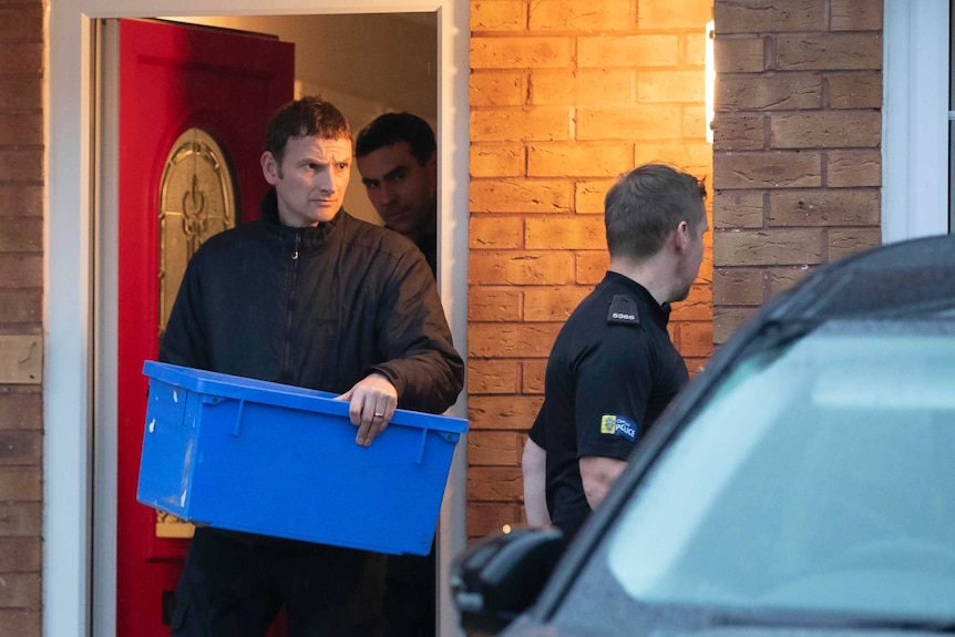 Three policemen walk out of a house one of them is carrying a blue plastic box