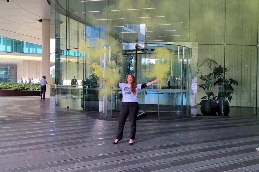 A woman stands outside an office building, releasing green gas from vessels in each hand.
