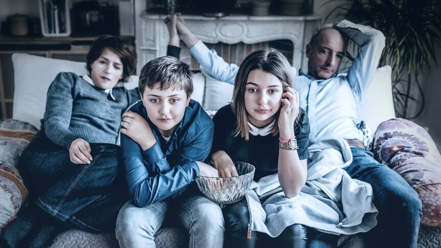 Family dressed in blue clothes sitting on couch, engrossed in television program. Two kids in foreground have a popcorn bowl.