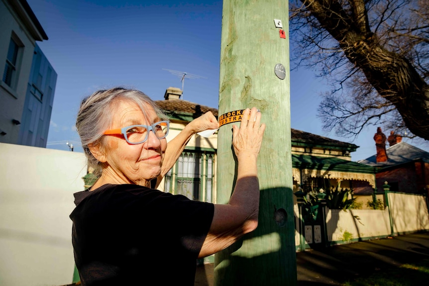 A grey-haired woman in a black tee puts a blockade australia sticker on a power pole