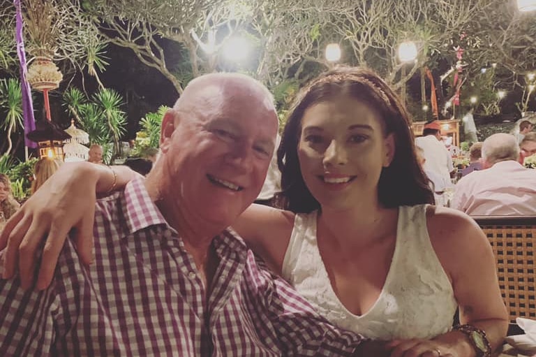 A middle-aged man in a red-and-white shirt smiles happily with a younger woman who is wearing a white top.