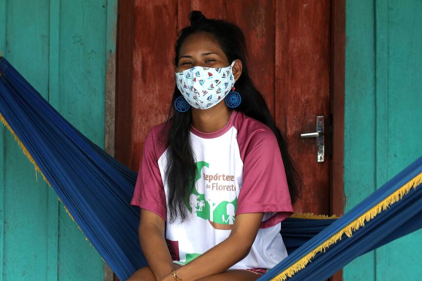 Dark haired young woman wearing pink shirt and white mask sits on a hammock against green building