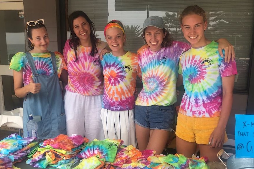 A group of girls wearing tie-dye shirts stand arm in arm in front of a table, covered with more tie-dye shirts