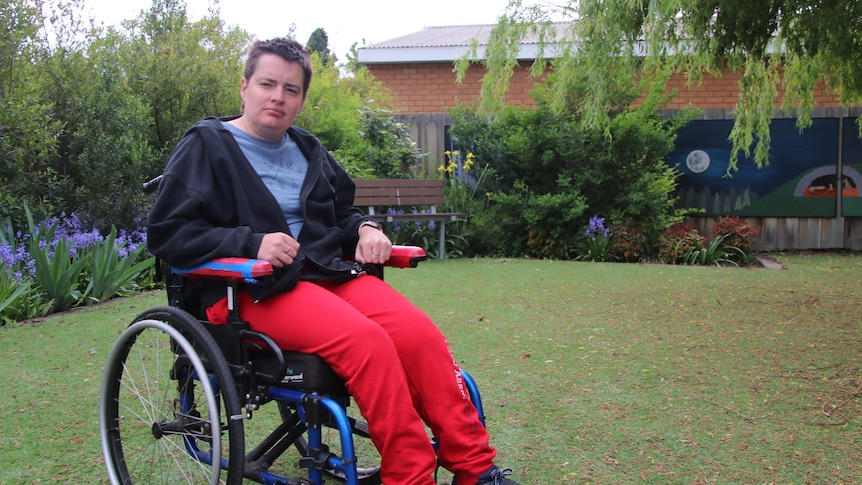 A serious young woman, short hair, red pants, blue top, dark blue jacket, sits in a wheelchair in a garden.
