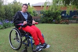 A serious young woman, short hair, red pants, blue top, dark blue jacket, sits in a wheelchair in a garden.