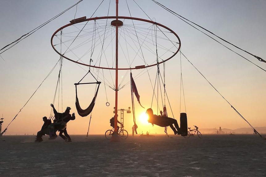 Revellers on a structure built with hammocks and swings in silhouette with the sun.