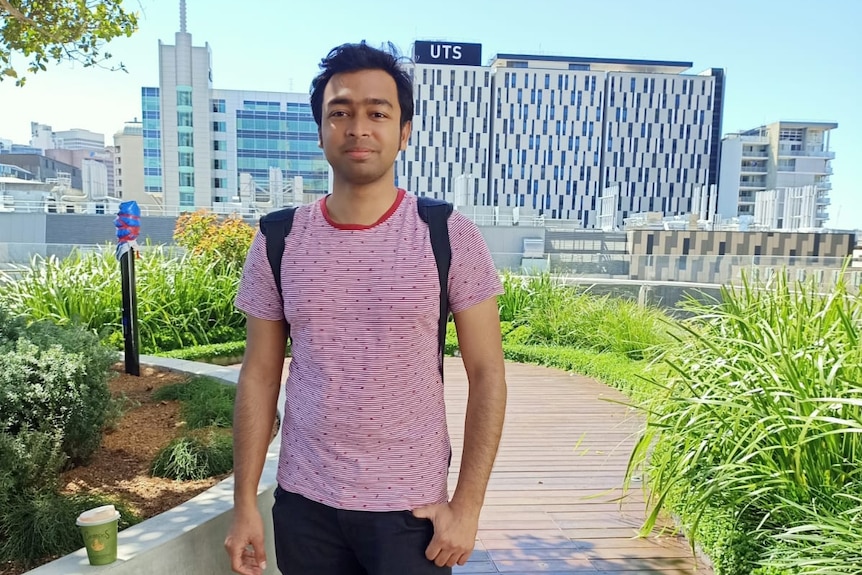 A man in a striped red t-shirt and black pants wearing a backpack stands in a garden with the UTS building behind him.