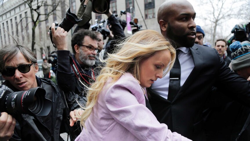 Stormy Daniels walks through a media pack with her head down, with the help of a man in a suit.