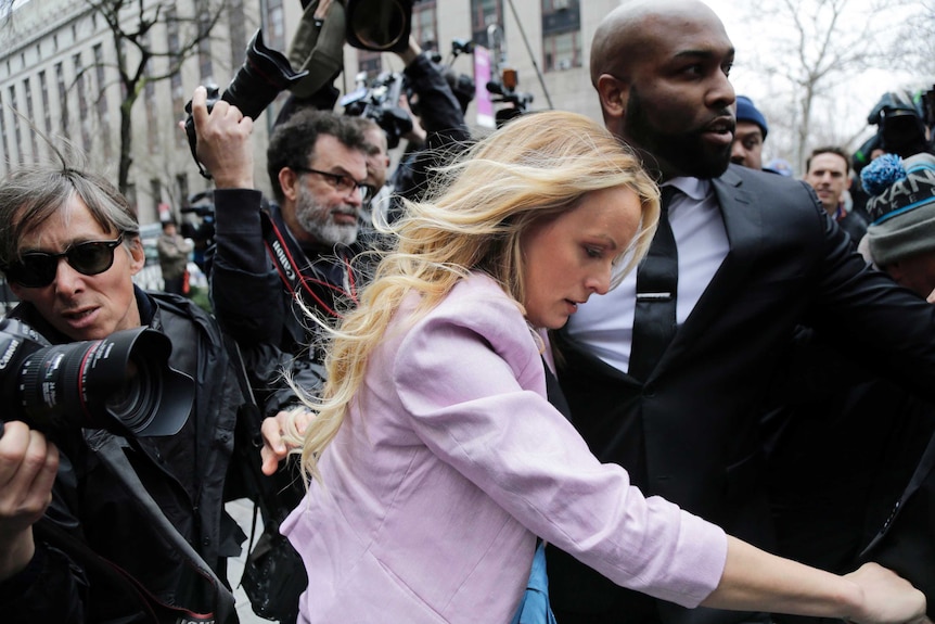 Stormy Daniels walks through a media pack with her head down, with the help of a man in a suit.