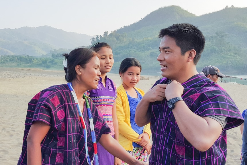 A young Asian man surrounded by women on a beach, all in traditional Burmese dress