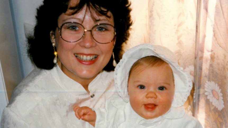 A woman dressed in white holding a baby in christening clothes and smiling at the camera.