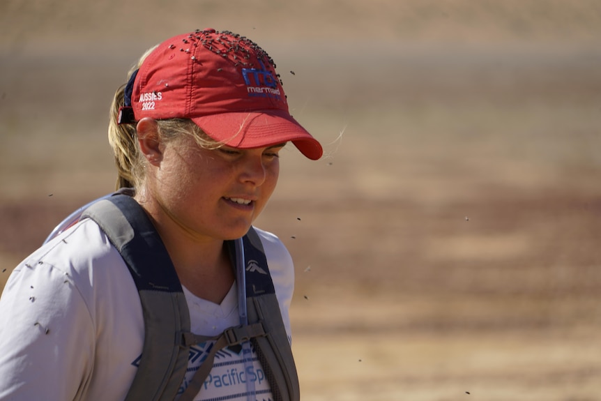 A female runner with a red cap covered in flies runs into the desert.