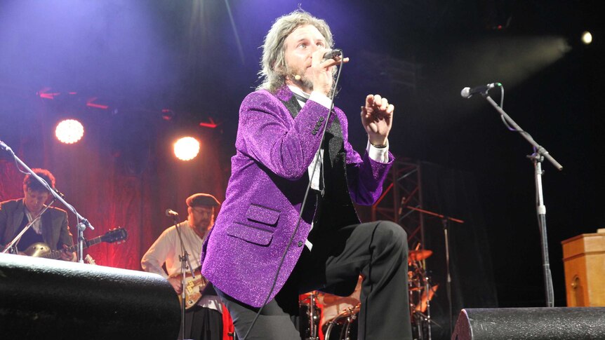 A man in a sparkly jacket sings in front of a band.