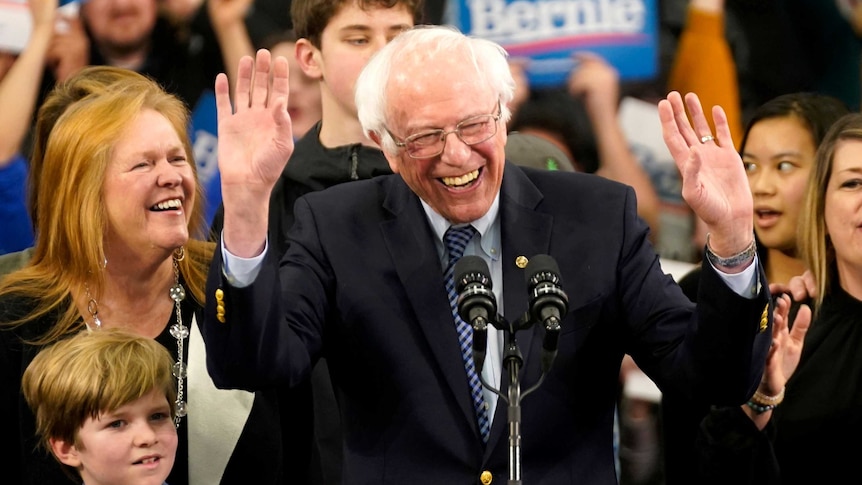 Bernie Sanders with his hands up smiling while standing at a podium