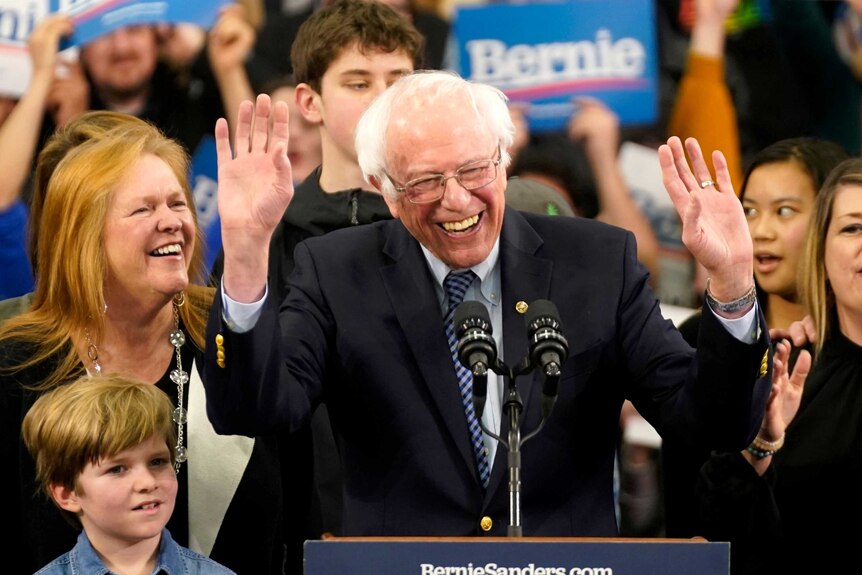 Bernie Sanders with his hands up smiling while standing at a podium