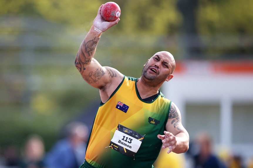 A man in a green and gold singlet hurls a heavy red ball through the air