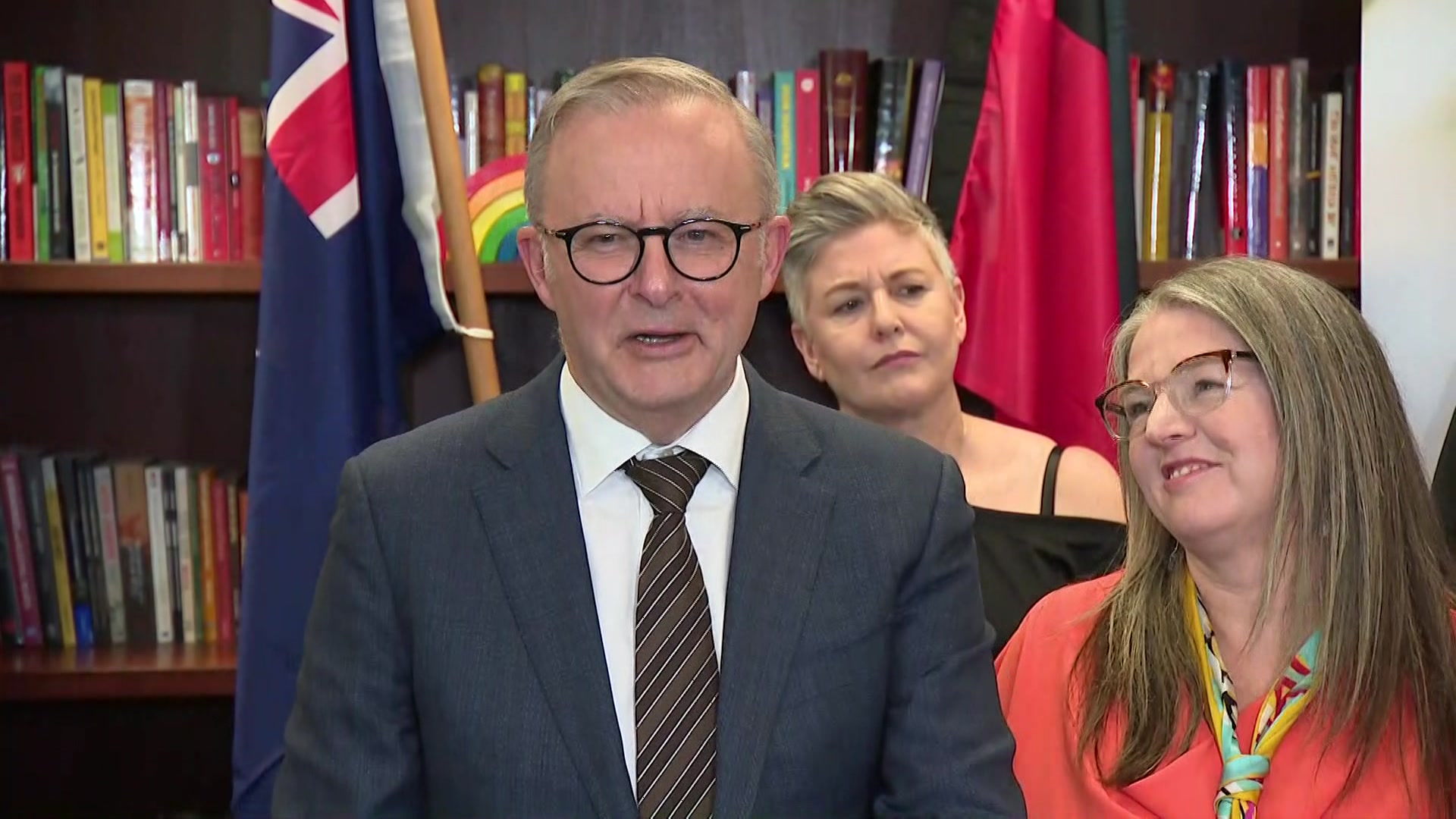 A man in a suit smiles as he speaks while two women watch on.