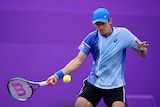 Tennis player Alex de Minaur returns serve with a forehand, wearing a cap on his head, on a grass court