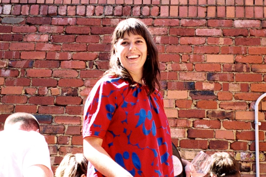 A woman wearing a floral top stands in front of a brick wall.
