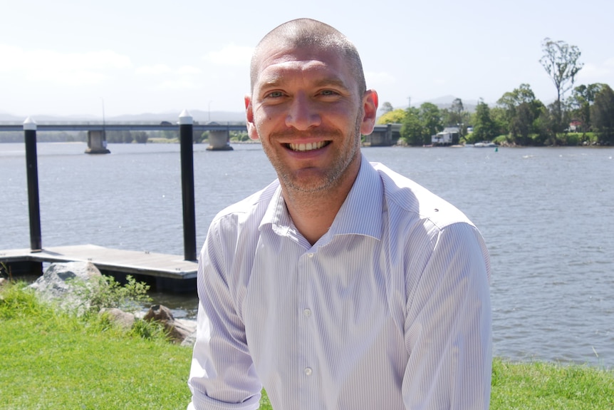 A man in a white t-shirt smiling by a river.