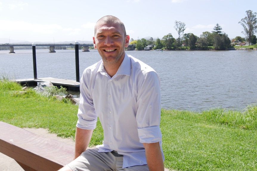 A man in a white t-shirt smiling by a river.