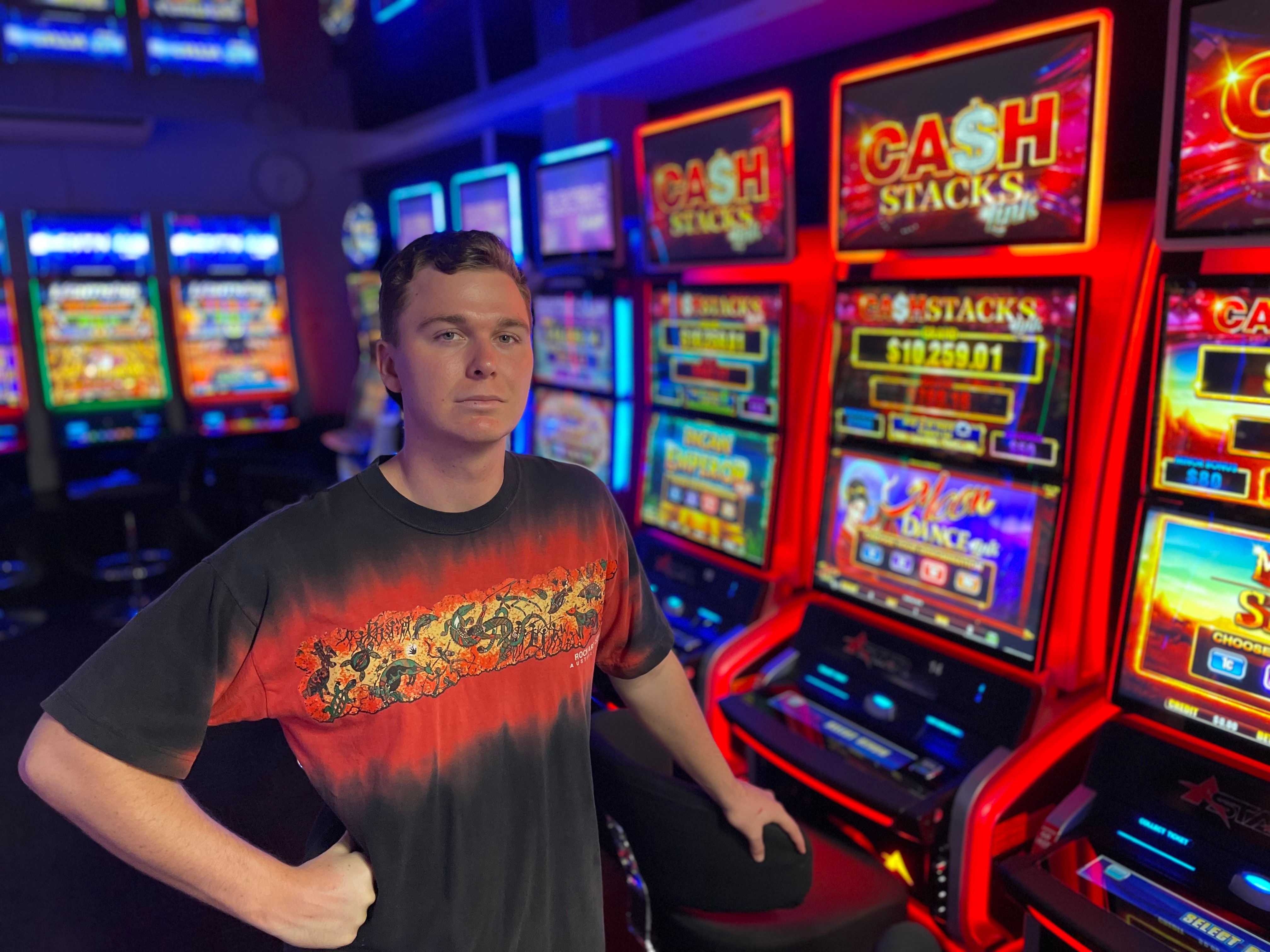 A young man with mousy hair and blue eyes stands in front of a pokies machine and frowns at the canberra 