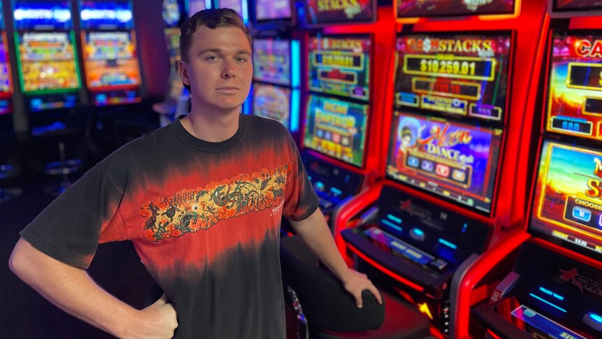 A young man with mousy hair and blue eyes stands in front of a pokies machine and frowns at the canberra 