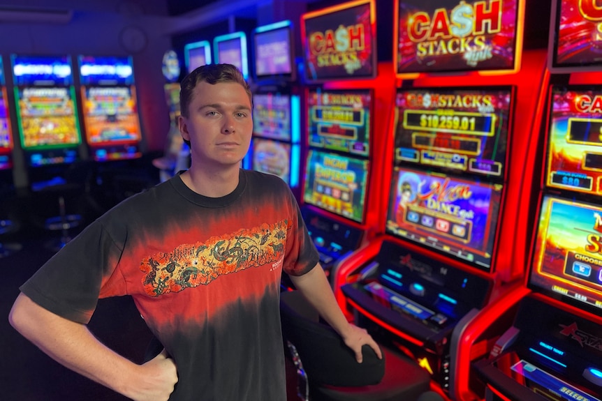 A young man with mousy hair and blue eyes stands in front of a pokies machine and frowns at the canberra 