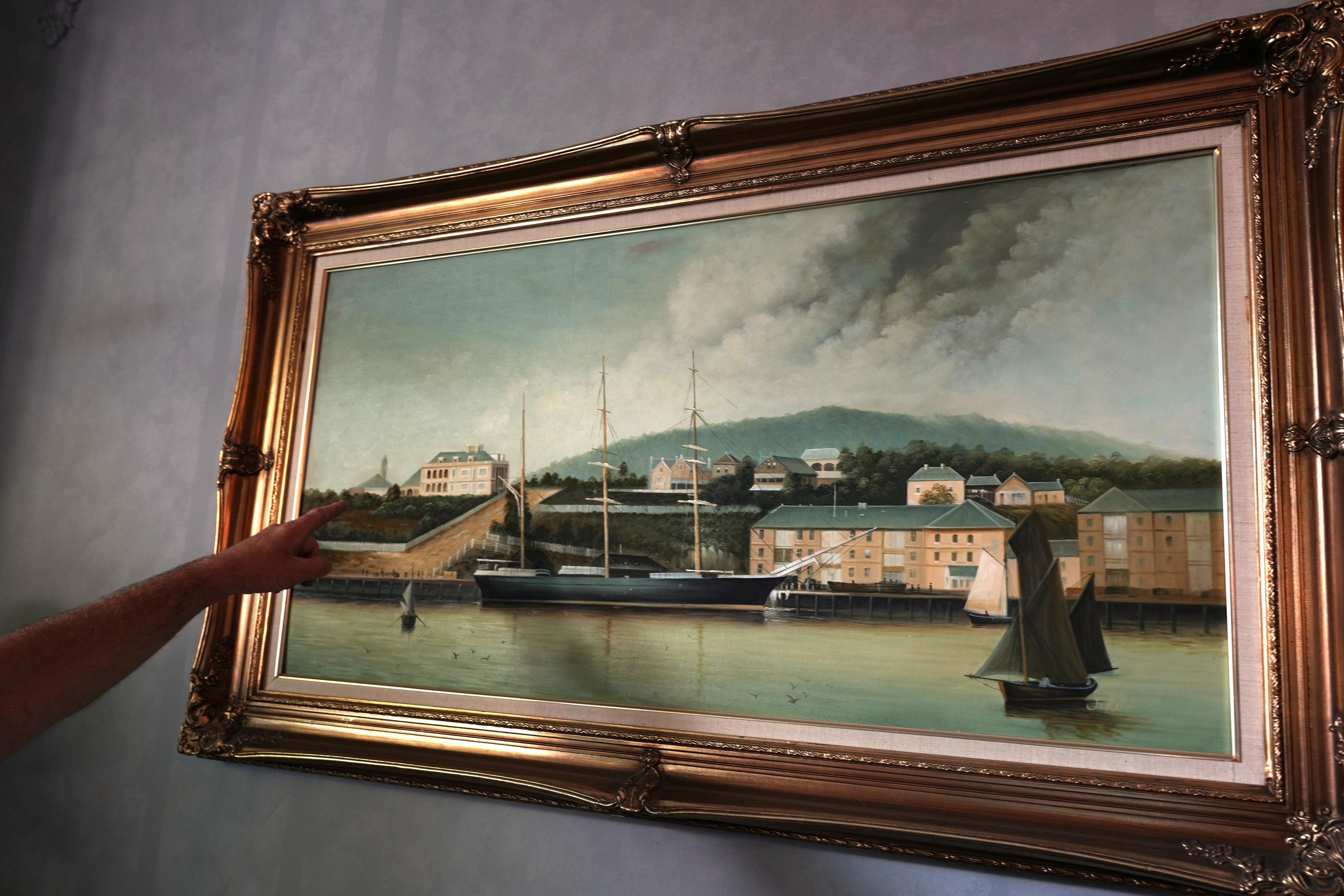 Un cuadro del puerto cuelga de una pared gris, con la mano de una persona señalando un detalle fuera de cámara.