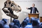 A man in a suit conducts an action behind a podium next to a large T-rex skull.
