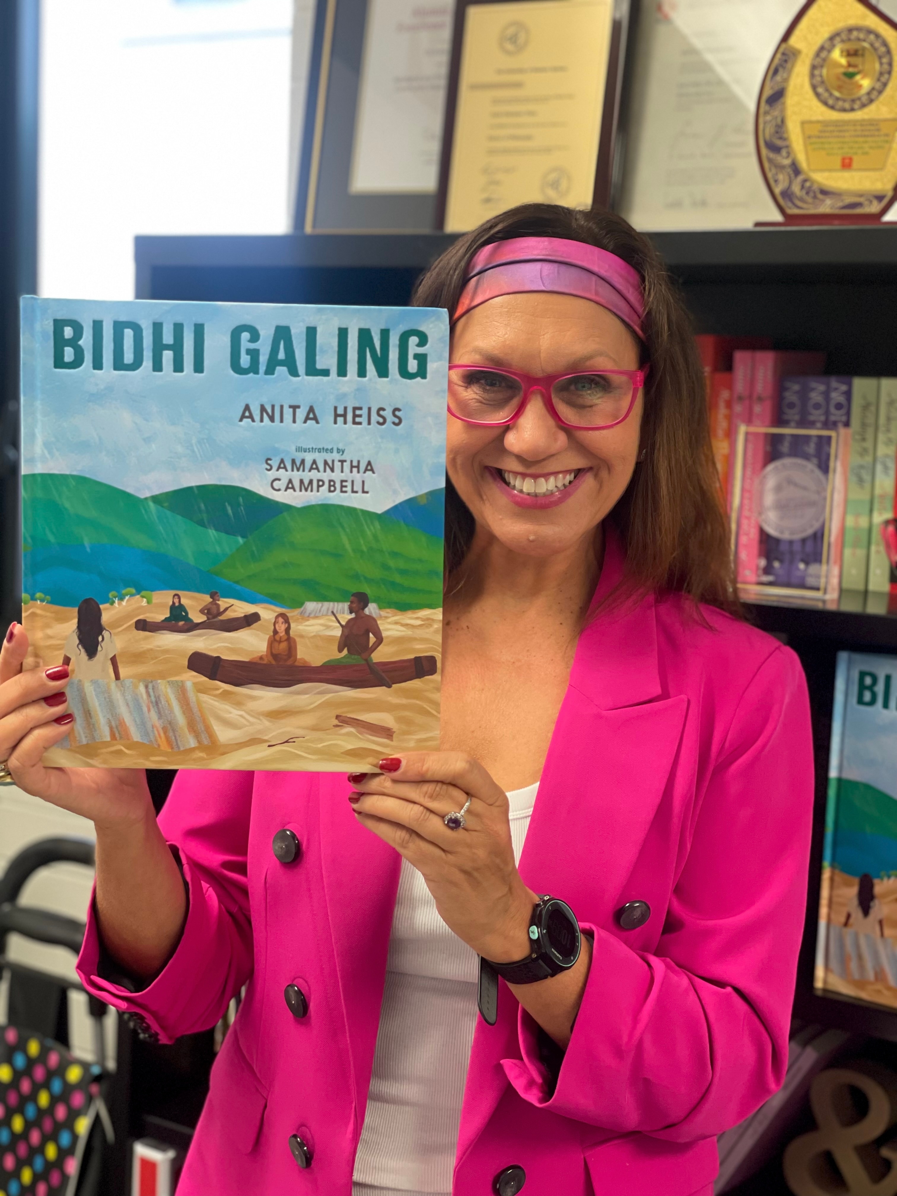 Anita Heiss dressed in a pink suit jacket, pink glasses and a pink and purple headband holding up one of her children's books