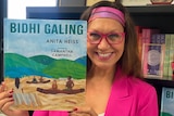 Anita Heiss dressed in a pink suit jacket, pink glasses and a pink and purple headband holding up one of her children's books