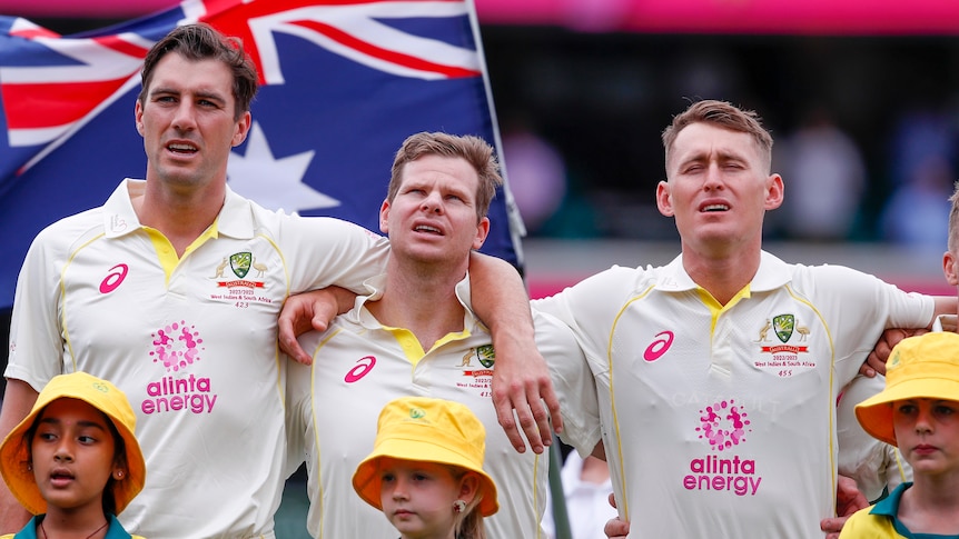 Three cricketers stand in front of an Australian flag and sing with their arms around each other.