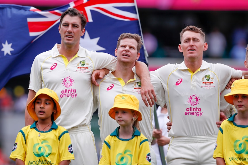Three cricketers stand in front of an Australian flag and sing with their arms around each other.