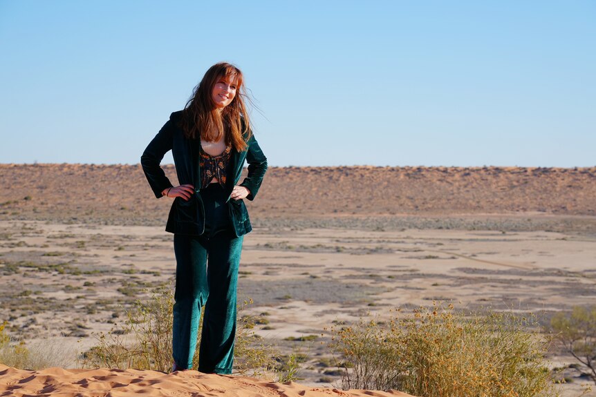A woman in a deep green suit stands atop a sandy dune hill