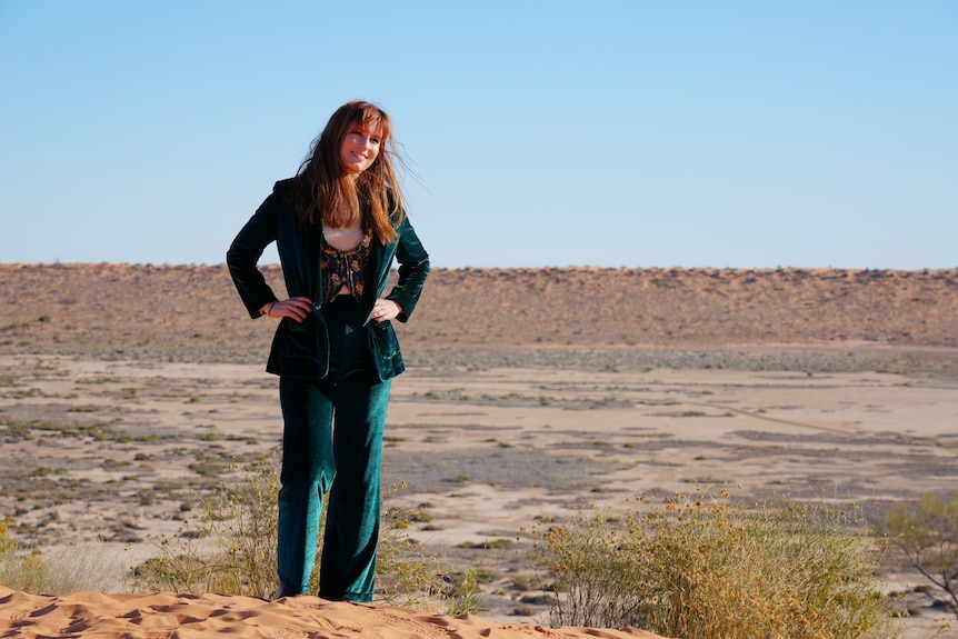 A woman in a deep green suit stands atop a sandy dune hill